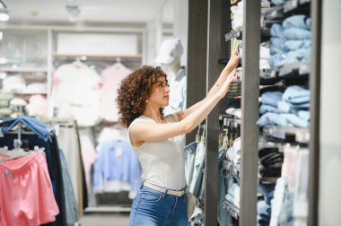 Şık bir mağazadan kıyafet almak için alışverişe çıkan zarif genç kadın renkli raflarda sergilenen elbiseleri inceliyor.