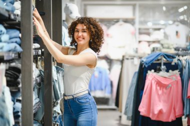 Gülümseyen genç bir kadın moda mağazasında kıyafet seçiyor, alışverişi seviyor ve yeni moda kıyafetler arıyor.