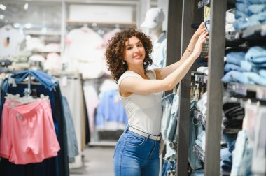 Kıvırcık saçlı gülümseyen kadın canlı bir moda mağazasında kıyafetleri karıştırıyor ve moda kıyafetleri seçme sürecinin tadını çıkarıyor.