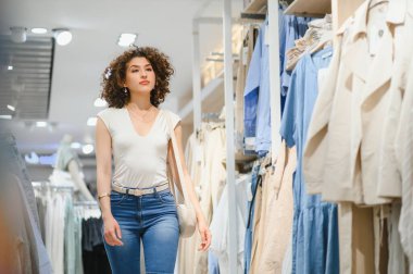 Hareketli bir moda mağazasında gezen şık genç bir kadın, kıyafetlerini ve aksesuarlarını seçerek mutlu mesut dolaşır.