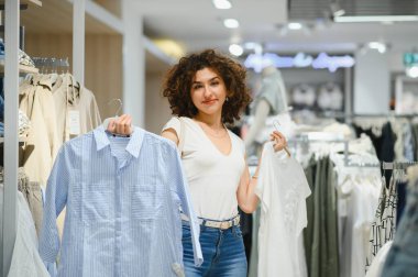 Giyim mağazasında alışveriş yaparken iki gömleği karşılaştıran genç bir kadın perakende terapi deneyiminin tadını çıkarıyor.