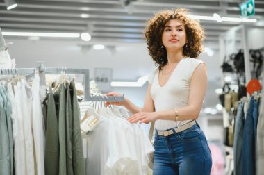 Moda mağazasında şık kıyafetler alırken canlı bir gömlek koleksiyonuna göz atan genç bir kadın.