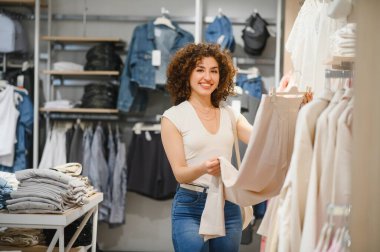 Mutlu bayan müşteri alışverişin tadını çıkarıyor, şık bej pantolonlar giyiyor ve canlı bir giyim mağazasında gülümsüyor.