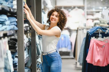 Gülümseyen genç bir kadın moda mağazasında kıyafet seçiyor, alışverişi seviyor ve yeni kıyafetler alıyor.