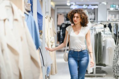 Genç bir kadın şık kıyafetlerini askılarda gezdirirken canlı bir moda mağazasında dolaşırken alışveriş deneyiminin tadını çıkarıyor.