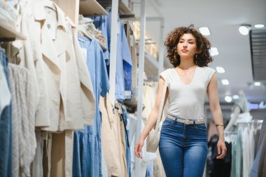 Genç bir kadın giyim mağazası koridorlarında geziniyor, şık kıyafetler arıyor ve alışveriş deneyiminin keyfini çıkarıyor.