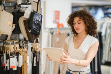 Moda mağazasından el çantası seçen şık genç bir kadın alışverişin tadını çıkarıyor ve yeni aksesuarlar arıyor.