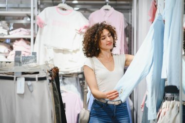 Kıyafet alışverişi yapan şık genç bir kadın, şık bir moda mağazasında dolaşırken canlı bir mavi gömleği zevkle inceliyor.