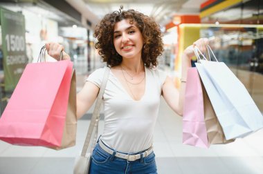 Alışveriş merkezinde eğlenen gülümseyen genç bir kadın, şık alışverişlerle dolu renkli alışveriş çantaları taşıyor.