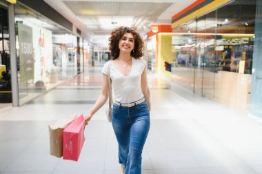 Neşeli, modern bir alışveriş merkezinde alışveriş çılgınlığının keyfini çıkaran mutlu müşteri, elinde çantalarla şık koridorlarda yürürken gülümsüyor.