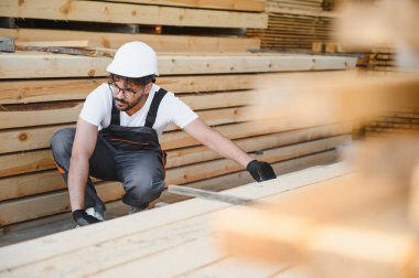Marangoz güvenlik kaskı takıyor ve bir depoda ahşap kalasları inceliyor, marangozluk ve kalite kontrolü konusunda uzmanlık gösteriyor.