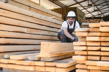 Marangoz güvenlik kaskı takıyor kereste deposunda ahşap kalaslar düzenliyor, marangozluk uzmanlığı ve endüstriyel işyeri sergiliyor.