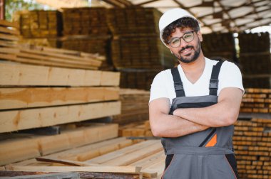 Güvenilir marangoz kaskı takıyor ve kereste deposunda çapraz kollarla gülümsüyor. Kereste endüstrisinde profesyonellik ve uzmanlık gösteriyor.