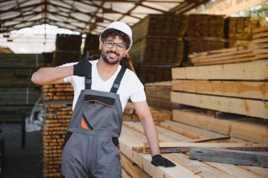 Gülümseyen marangoz iş tulumu giyer ve ambardaki kereste yığınlarının yanında dikilirken başparmak hareketi yapar.
