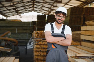 Kereste deposunda çapraz kollarla duran kendinden emin bir ahşap işçisinin portresi kereste endüstrisinde uzmanlık ve gurur gösteriyor.