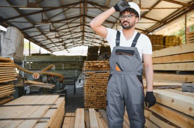 Yorgun marangoz, kereste deposunda çalışırken alnından ter siliyor. Etrafı tahta yığınlarıyla çevrili.