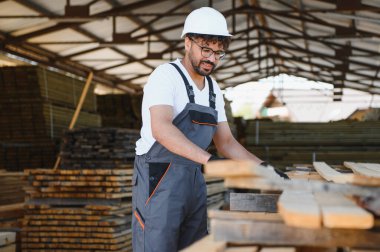 Marangoz kereste deposundaki kereste kalaslarını seçiyor, koruyucu ekipmanlarla işyeri güvenliğini gösteriyor.