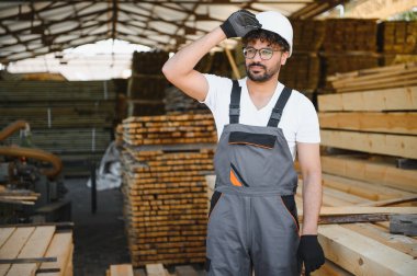 Marangoz kereste deposunda çalışırken şapkasını düzeltirken yorgun düşmüş. Etrafı tahta yığınlarıyla çevrili.