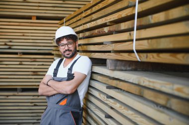 Çapraz kolları olan kendine güvenen marangoz bir depodaki kereste yığınına yaslanıyor, marangozluk endüstrisinde uzmanlık ve profesyonellik gösteriyor.