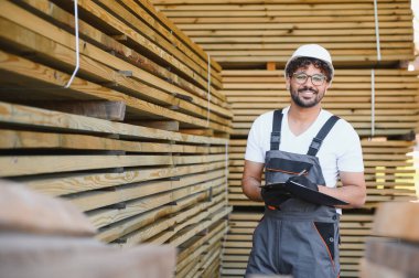 Marangoz başlık ve tulum giyer. Depodaki ahşap kalasları incelerken panoya not alır.