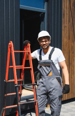 İnşaat işçisi, işçi tulum giyiyor ve inşaat sahasında merdivene yaslanmış, çalışmaya hazır.