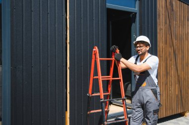 İnşaat işçisi, prefabrik bir ev inşa ederken elinde merdiven tutuyor, modern konut gelişimine kendini adamış.