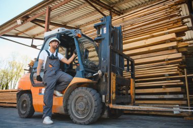 Depo işçisi mola veriyor kereste deposundaki kereste yığınlarının yanında bir forkliftte oturuyor.