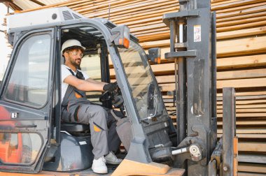 Güvenlik kaskı takan ve iş tulumu giyen bir depo işçisi kereste deposunda forklift kullanıyor, ahşap tahtalar taşıyor.