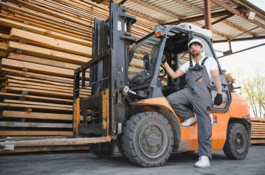 Depoda işçi işçi tulum giyiyor ve forklift kullanıyor. Açık hava deposundaki kereste yığınlarının yanında.