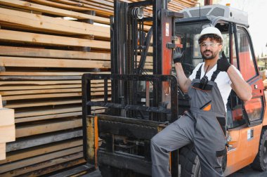 Depo işçisi, kereste yığınının yanında bir forkliftte otururken, iş memnuniyetini ve verimliliğini gösterirken baş parmağını kaldırıyor.