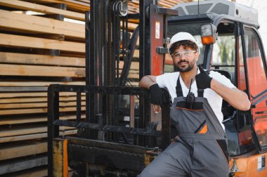 Depo çalışanı iş tulumu ve başlık takıyor. Kereste deposundaki kereste yığınlarının yanında bir forklifte yaslanırken başparmak kaldırıyor.
