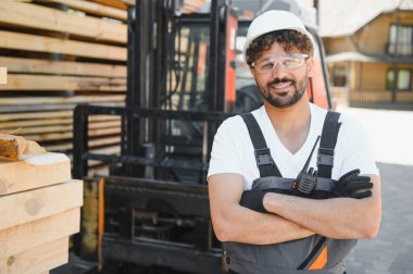Kereste deposunda kollarını çapraz bağlamış, güvenlik kıyafeti giymiş gülümseyen bir Arap kereste işçisinin portresi.