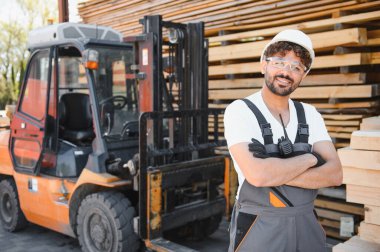 Güvenlik gözlüğü takan gülümseyen Arap depo çalışanı portresi ve forklift ve kereste yığınlarının yanında çapraz kollu şapka.