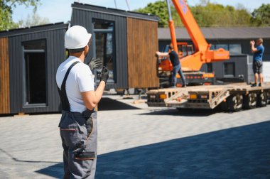 İnşaat işçisi telsiz kullanıyor, vinç yönetiyor prefabrik modüler bir evi kamyona yüklüyor, etkin inşaat çalışmalarını gösteriyor.
