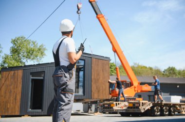 İnşaat işçisi telsiz kullanıyor, vinç kaldırmayı koordine ediyor prefabrik modüler evi kamyona yüklüyor, etkin bina ulaşımı sergiliyor.