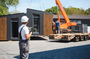 İnşaat alanında vinç ve kamyon kullanarak prefabrik evin taşınmasını kontrol eden mühendis.