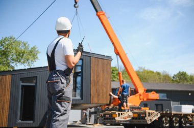 İnşaat işçisi telsiz kullanıyor, vinç kaldırmayı denetliyor prefabrik modüler evi kamyona yüklüyor, verimli konut taşımacılığı için yenilikçi yapı teknolojisi