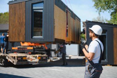 Beyaz başlık ve eldiven takan mühendis, prefabrik modüler bir evin güvenli ve verimli bir şekilde nakledilmesini sağlayan bir kamyona yüklenmesini denetliyor.
