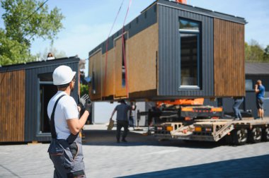 Mühendis prefabrik modüler bir evin bir kamyona yüklenmesini denetliyor, varış noktasına verimli ve güvenli bir şekilde ulaşmasını sağlıyor.