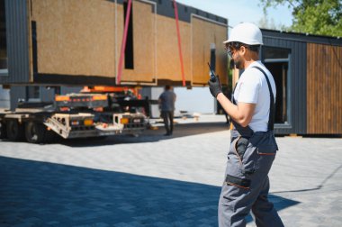 İnşaat işçisi telsiz kullanıyor, modüler evi koordine ediyor kamyonete yüklüyor, şantiyeye güvenli ve verimli ulaşım sağlıyor.