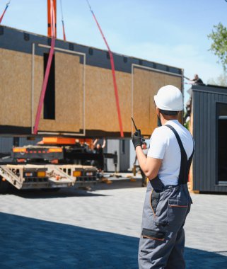 İnşaat işçisi telsiz kullanıyor, vinç yönetiyor prefabrik bir ev modülünü kamyona kaldırıyor, güvenli ve verimli bir ulaşım sağlıyor.