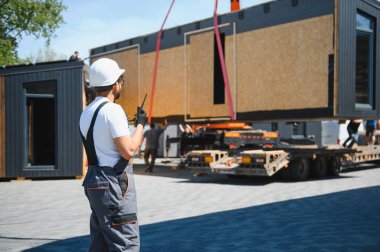 İnşaat işçisi telsiz kullanıyor, prefabrik modüler bir evin kamyonete yüklenmesini koordine ediyor, güvenli ve verimli bir ulaşım sağlıyor.