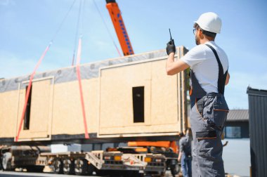 İnşaat işçisi telsiz kullanıyor, vinç kaldırmayı koordine ediyor prefabrik ev bölümünü kamyona yüklüyor, modüler yapının verimliliğini gösteriyor.