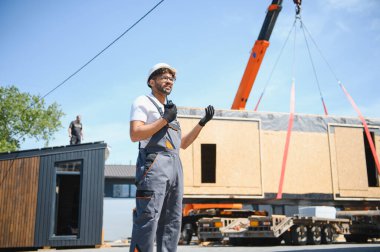 İnşaat işçisi telsiz kullanıyor ve vinç kaldırmayı denetliyor prefabrik modüler evi kamyona yüklüyor, yenilikçi inşaat yöntemi