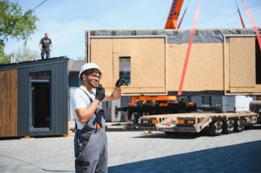 İnşaat işçisi telsiz kullanıyor ve prefabrik modüler ev bölümünün kamyonete yerleştirilmesini koordine ederken işaret ediyor.