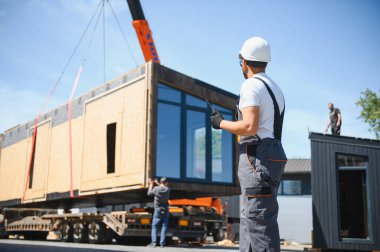 İnşaat işçisi telsiz kullanıyor. İnşaat alanındaki kamyona prefabrik modüler ev yüklemesini denetliyor.