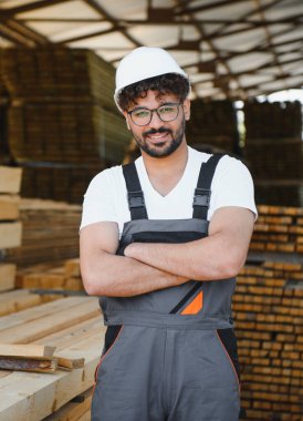 Kendine güvenen bir ahşap işçisinin portresi çapraz kollarla gülümseyen, kereste deposunda duran, kereste endüstrisinde uzmanlık ve profesyonellik gösteren