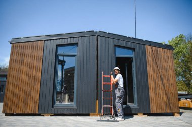 İnşaat işçisi bir merdiven tutuyor ve prefabrik modüler bir evin kamyonun üzerine kaldırılmasını denetliyor, yenilikçi ve sürdürülebilir yapı çözümü.