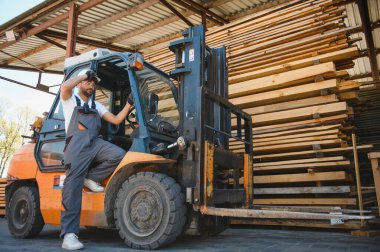 Depo işçisi açık hava depolama alanındaki kereste yığınlarının yanında forklift kullanmaya ara veriyor.