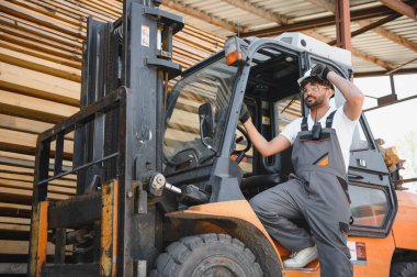 Depo işçisi tulum giyiyor ve forklift kullanıyor. Büyük bir sanayi deposunda kereste düzenliyor.